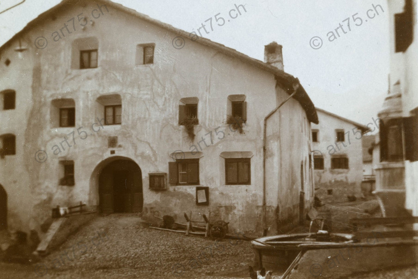 Historische Fotografie eines Engadinerhauses in Ardez im Unterengadin mit Brunnen im Vordergrund, aufgenommen um 1920.