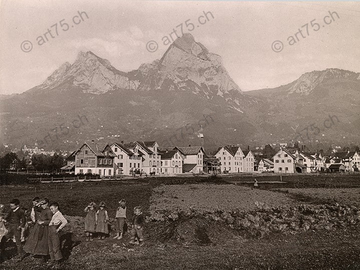 Historische Aufnahme von Ibach bei Schwyz um 1900 mit spielenden Kindern, Häusern und dem markanten Doppelgipfel der Mythen im Hintergrund.